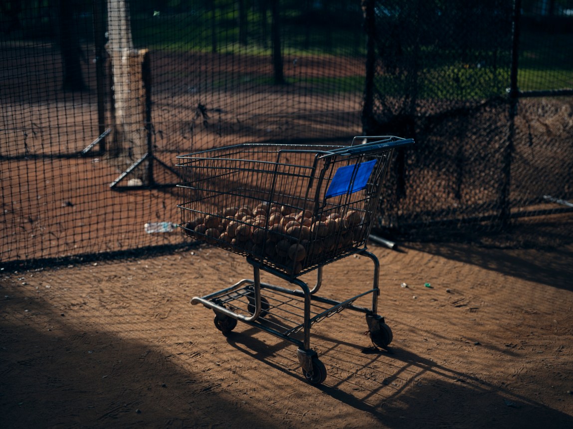 A wire shopping cart filled with worn baseballs sits on a dirt infield behind a protective batting net.