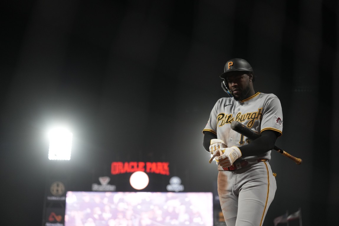 A man in a Pittsburgh uniform and helmet has a baseball bat under his arm. He is adjusting his gloves. Behind him are the bright lights of a stadium and a dark sky.