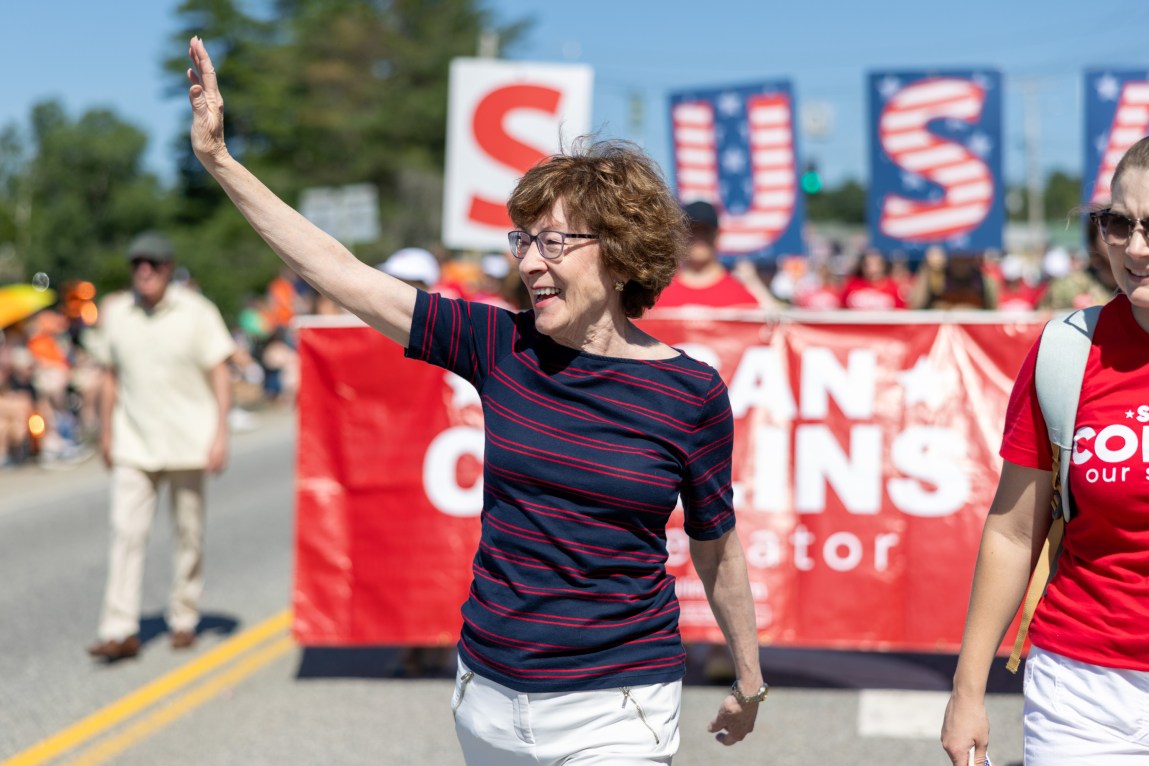 A woman in a blue T-shirt with red stripes waves to the crowd as she walks in front of a red banner during a parade.