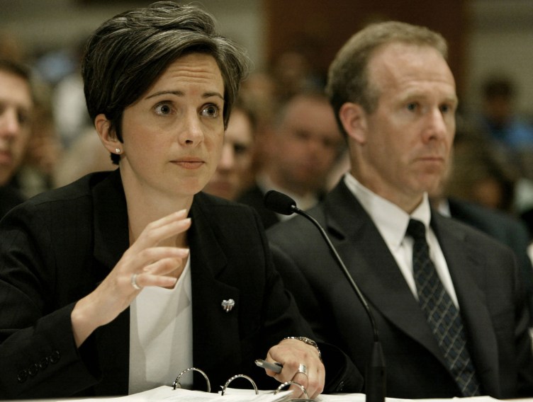 A woman in business attire gestures with her hand as she speaks into a microphone. A man in a suit sits next to her, and numerous people look on in the background.