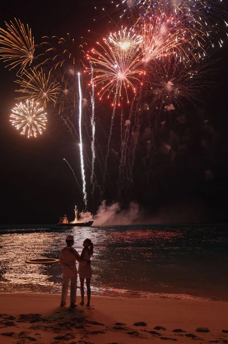 A man and woman stand on a beach as fireworks explode above the water.