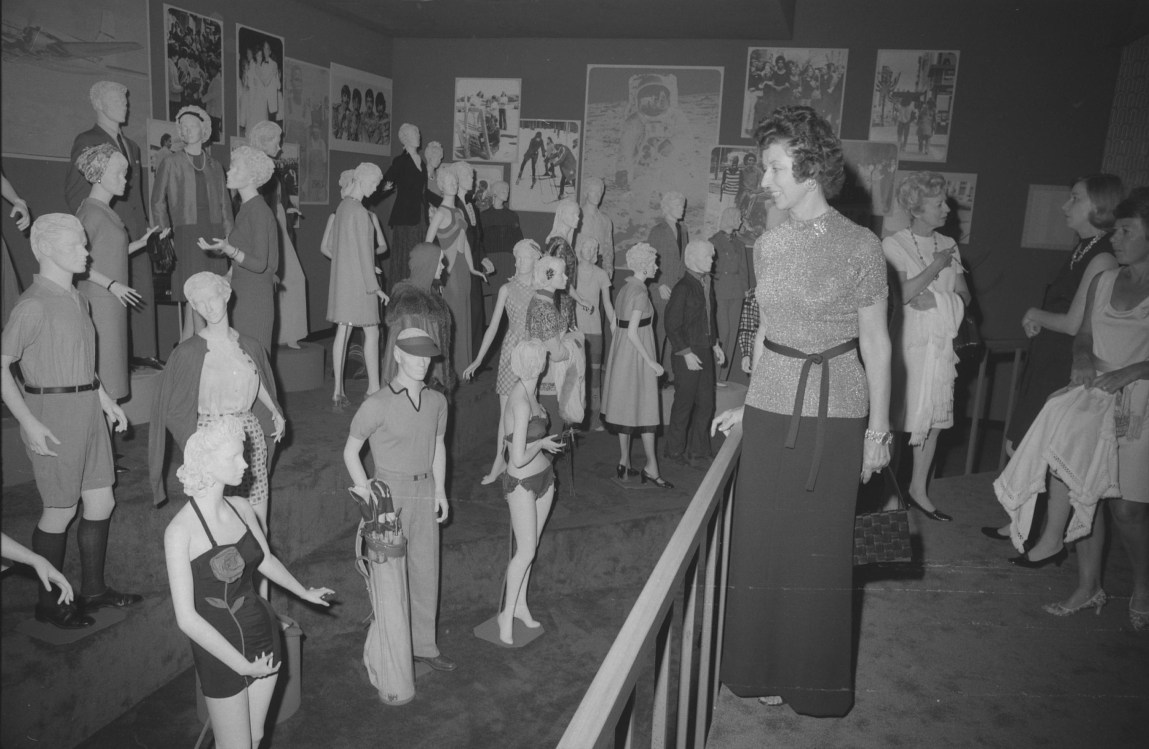 A black-and-white photo of women looking at an exhibit of mannequins in different outfits. 
