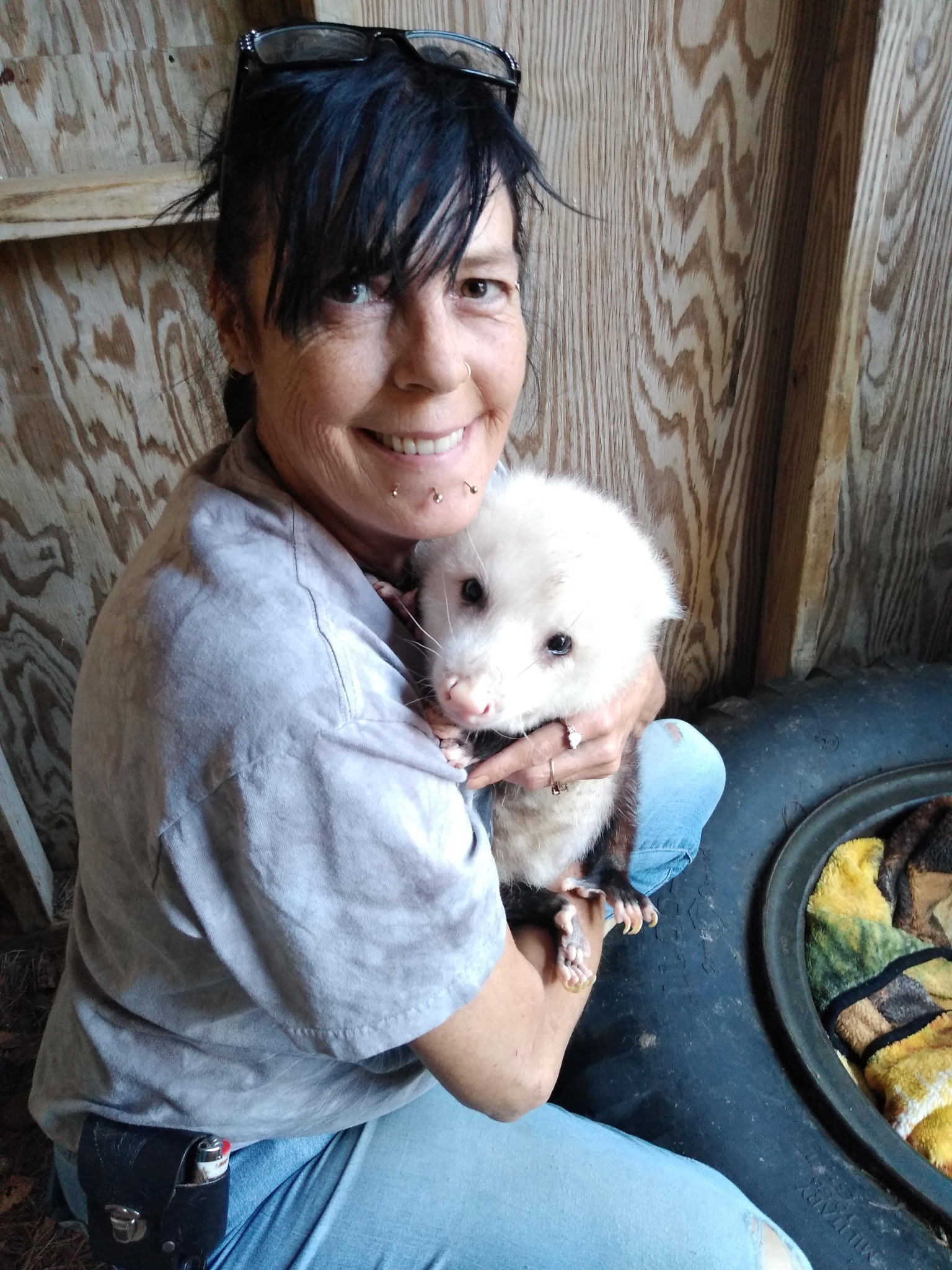 A woman with black bangs and chin piercings smiles and holds a white opossum. She is sitting in front of a wooden wall.