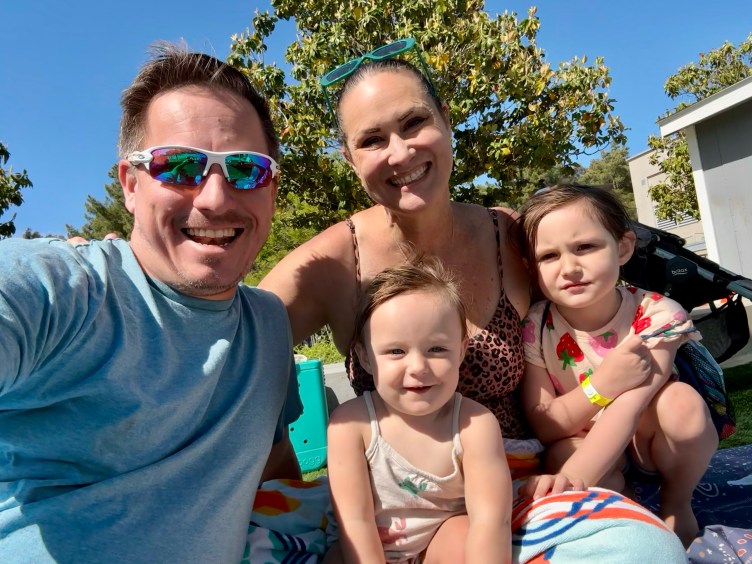 A smiling family of four taking a sunny outdoor selfie, featuring a man in white sunglasses, a woman, and two young girls sitting together.