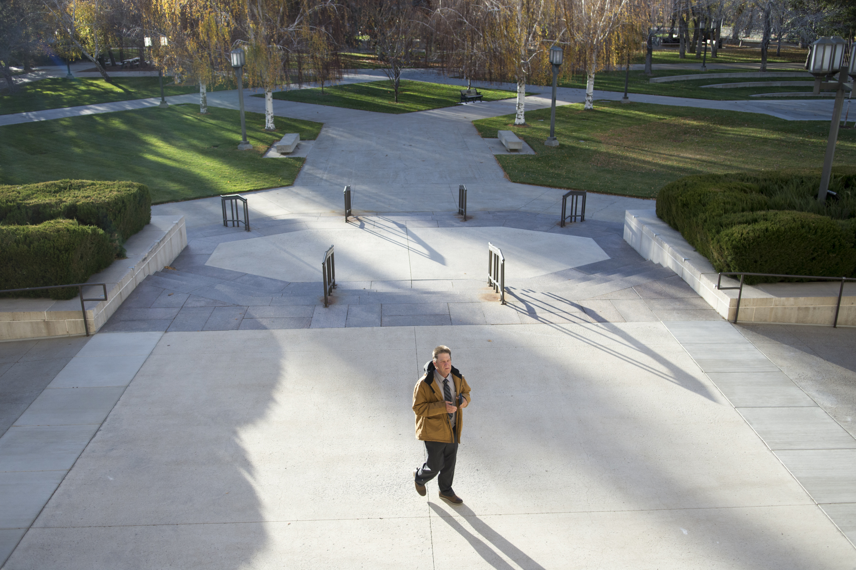An aerial view of a large staircase with manicured lawns and trees. A man wearing a mustard coat and gray tie stands in the center, with his shadow cast before him.