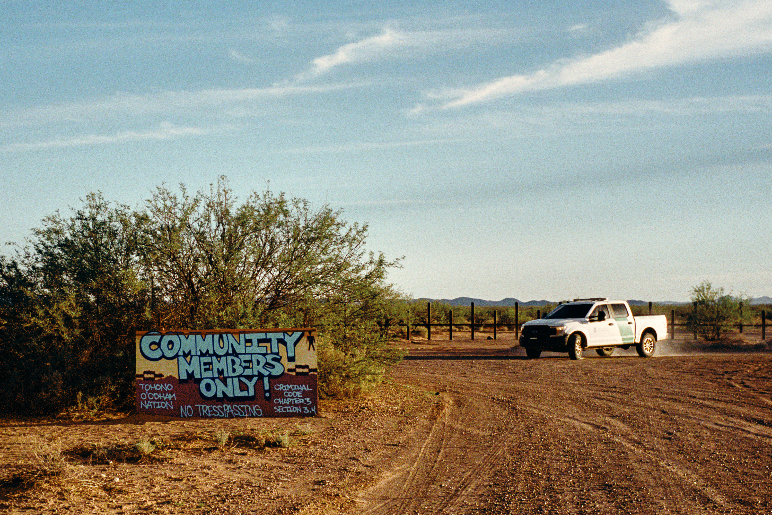 On the left, a red and yellow painted sign with graffiti-style white letters warns that “Community Members Only!” are allowed on the Tohono O’odham Nation. A white and green Border Patrol truck is driving on a dirt road on the right.