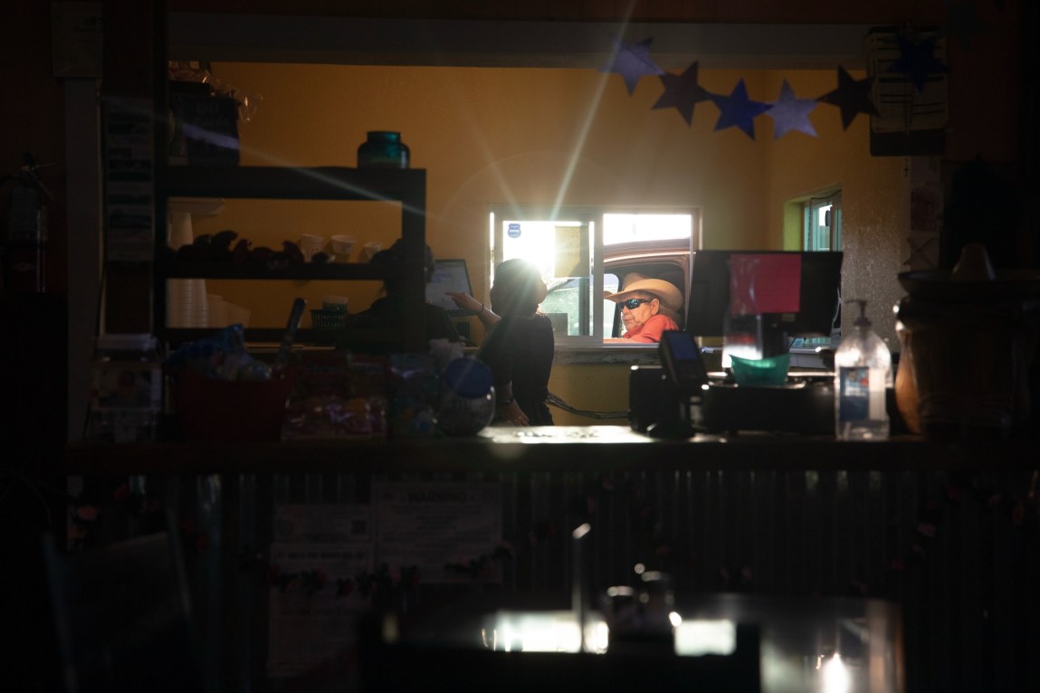 The dark interior of a Mexican restaurant is illuminated by light from a drive-thru window. A woman is working at a register by the window, where a customer wearing a cowboy hat has driven up.