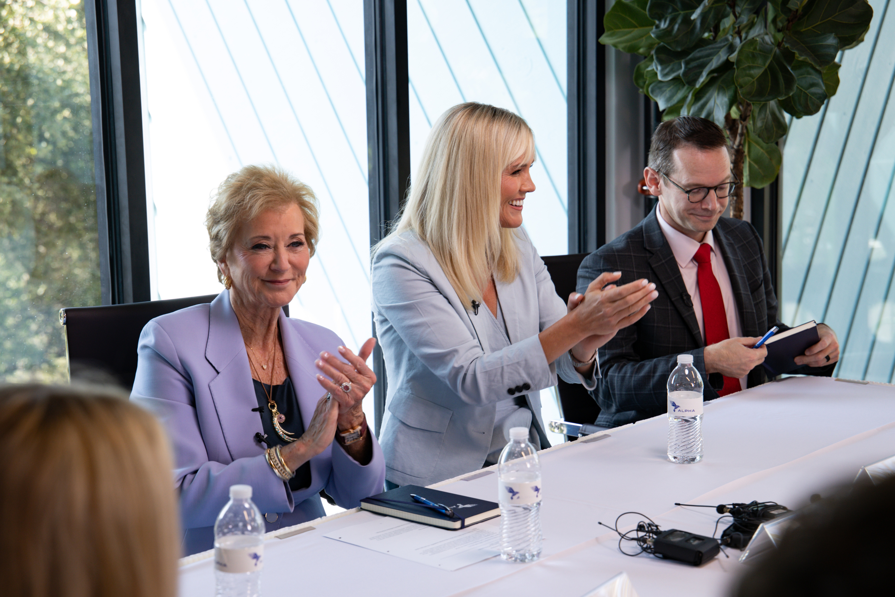 People wearing suits sit in office chairs around a white table in a room with glass windows. Two of the women are clapping and smiling, and one of the men looks down while holding a notebook.
