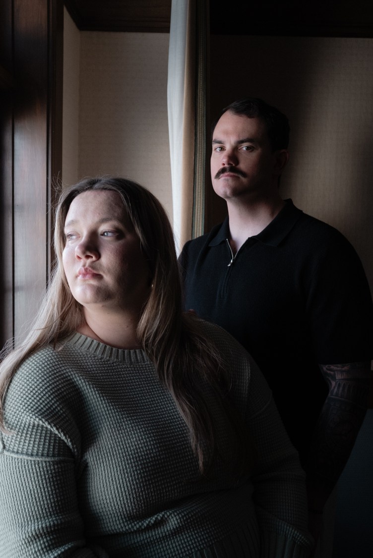 A portrait of a woman looking thoughtfully out a window while a man with a mustache stands in the background behind her.