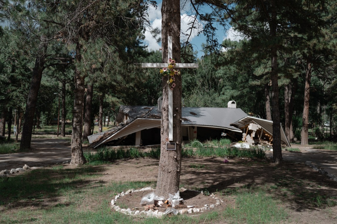 A wooden cross decorated with flowers stands attached to a tree in front of a collapsed building in a wooded area.