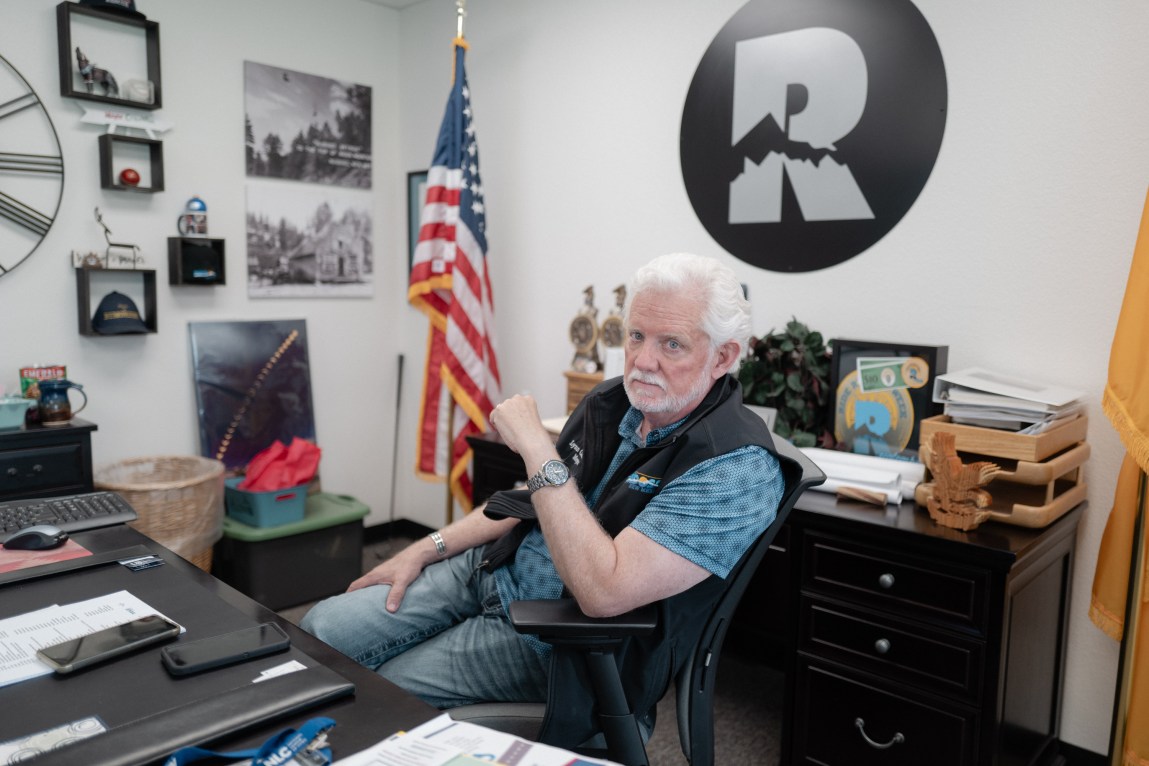 A man with white hair and a white beard sits at an office desk in front of an American flag and village logo, resting his arm on his chair while looking toward the camera.