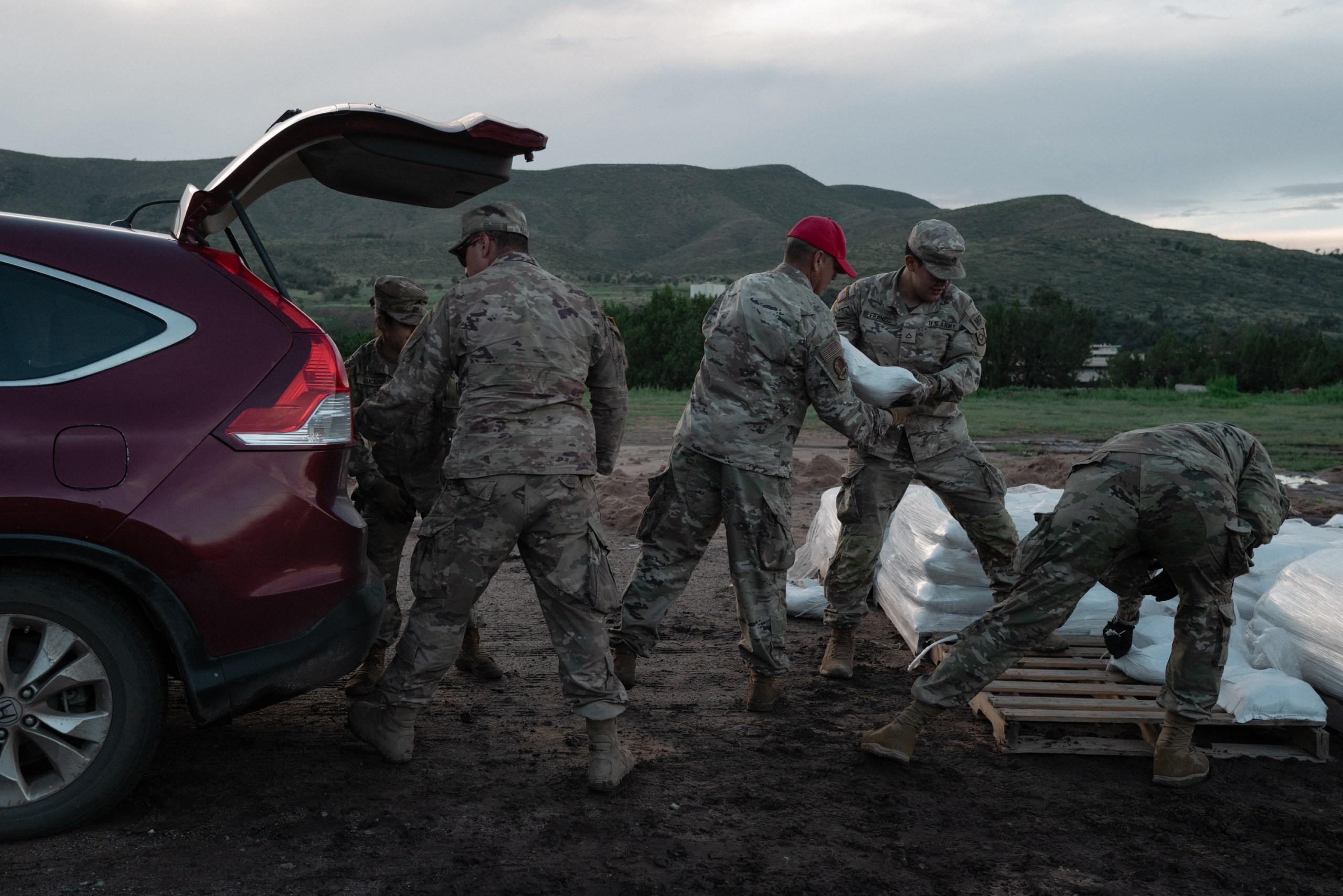 National Guard service members load white sandbags from wooden pallets into the open trunk of an SUV ahead of flood preparations.