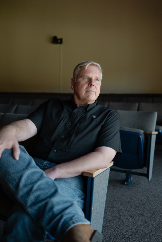 A man with short graying hair sits in an auditorium chair with his legs crossed, looking off to the side.