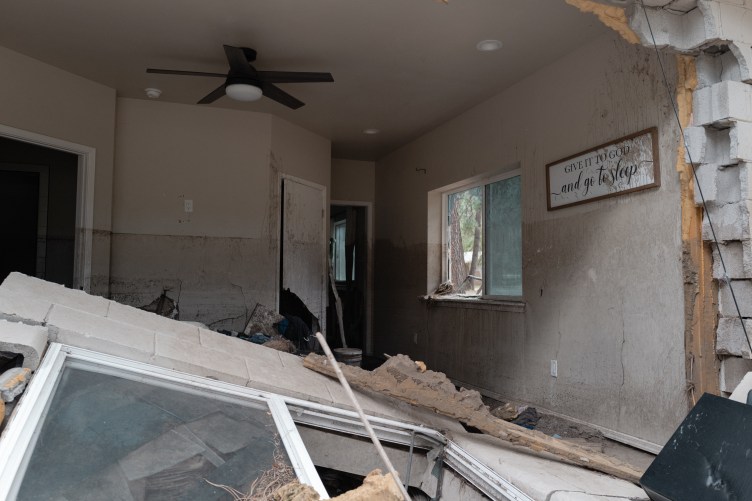 The interior of a home shows severe flood damage, high mud lines on the walls, collapsed cinder blocks and a framed sign reading “Give it to God and go to sleep.”