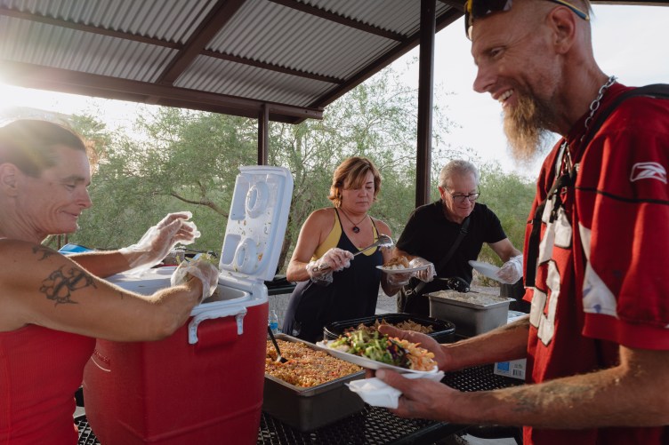 Volunteers serve warm food and sides to people standing in line under an outdoor shelter.
