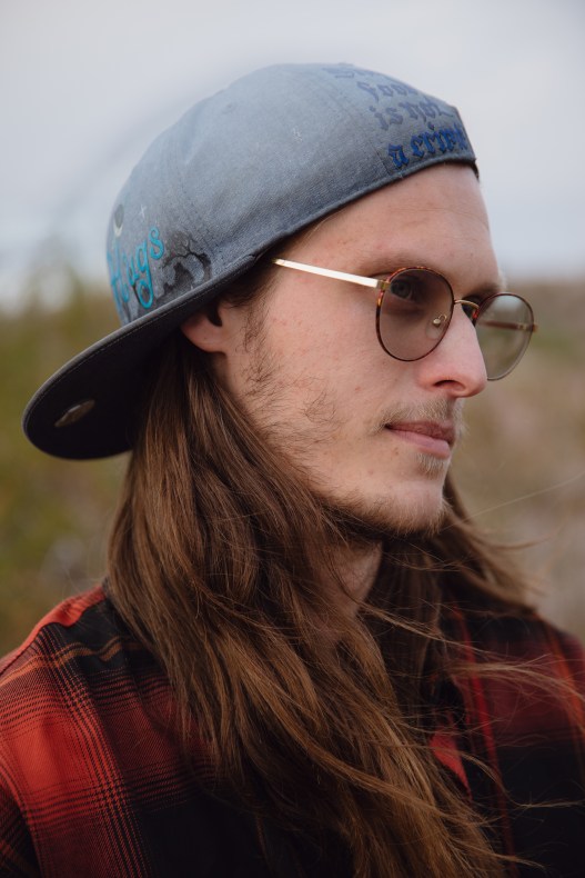 A close-up portrait of a man with long hair, round glasses and a gray baseball hat featuring handwritten text.