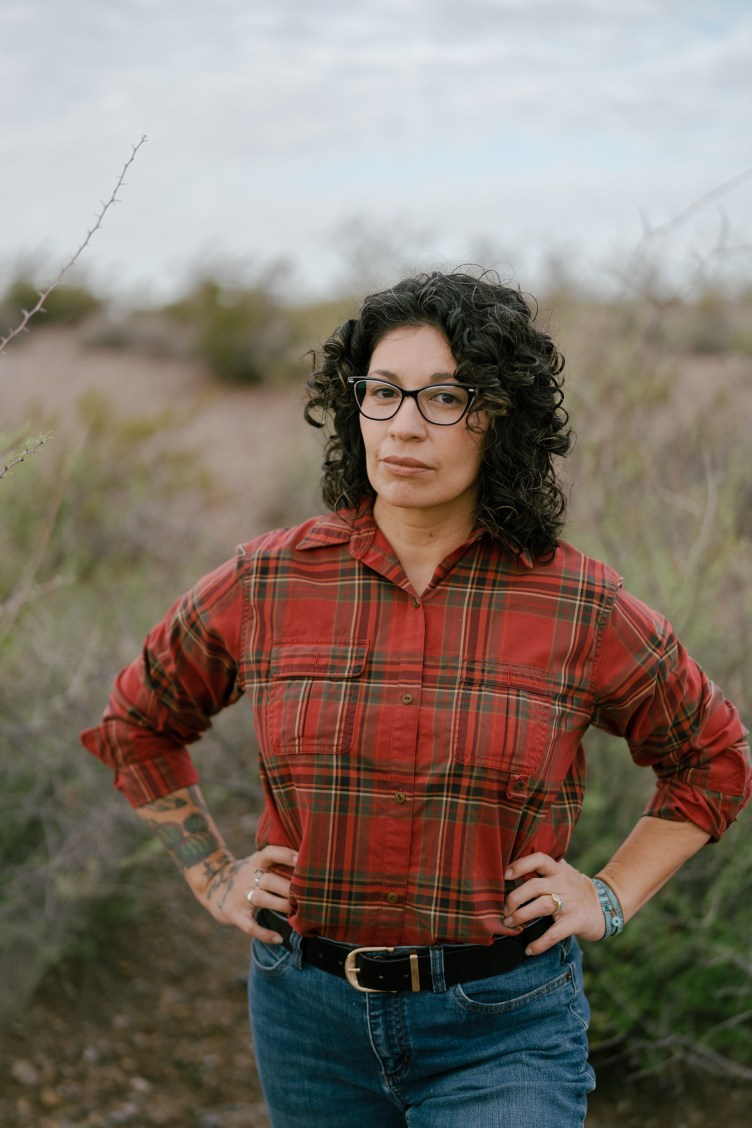 A portrait of a woman with dark curly hair and glasses, wearing a red plaid button-down shirt and blue jeans, standing outdoors in a desert landscape with her hands on her hips.