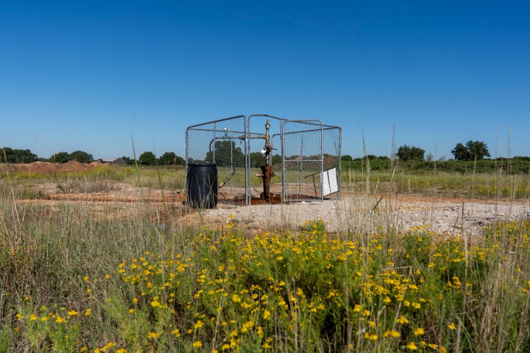 A structure made of pipes, with the lower ones deeply rusted, rises in a field with yellow flowers. Chain-link fencing encloses a small area around the pump.
