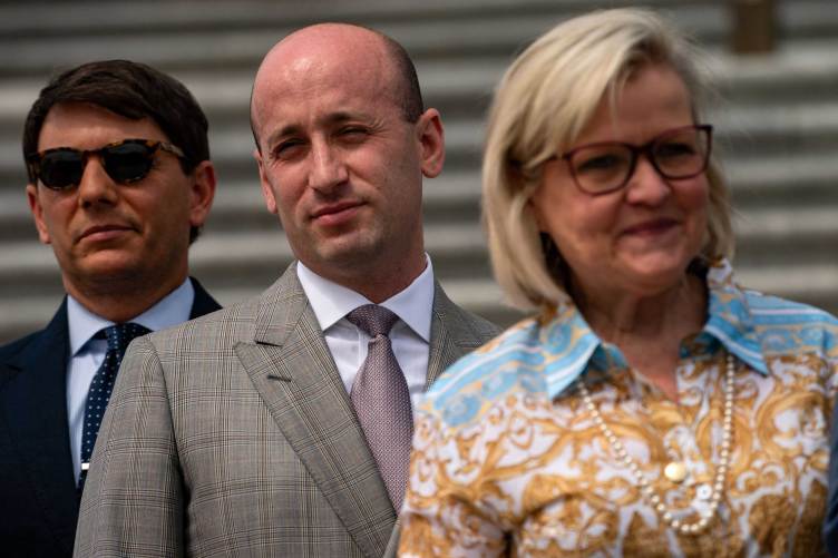 A man stands outdoors in a gray plaid suit and tie, positioned between a man wearing sunglasses on the left and a smiling Cleta Mitchell on the right.