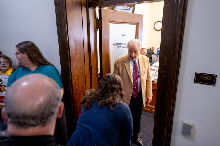 A man with white hair and glasses in a suit stands in a wooden doorway. A group of people stand outside the door.