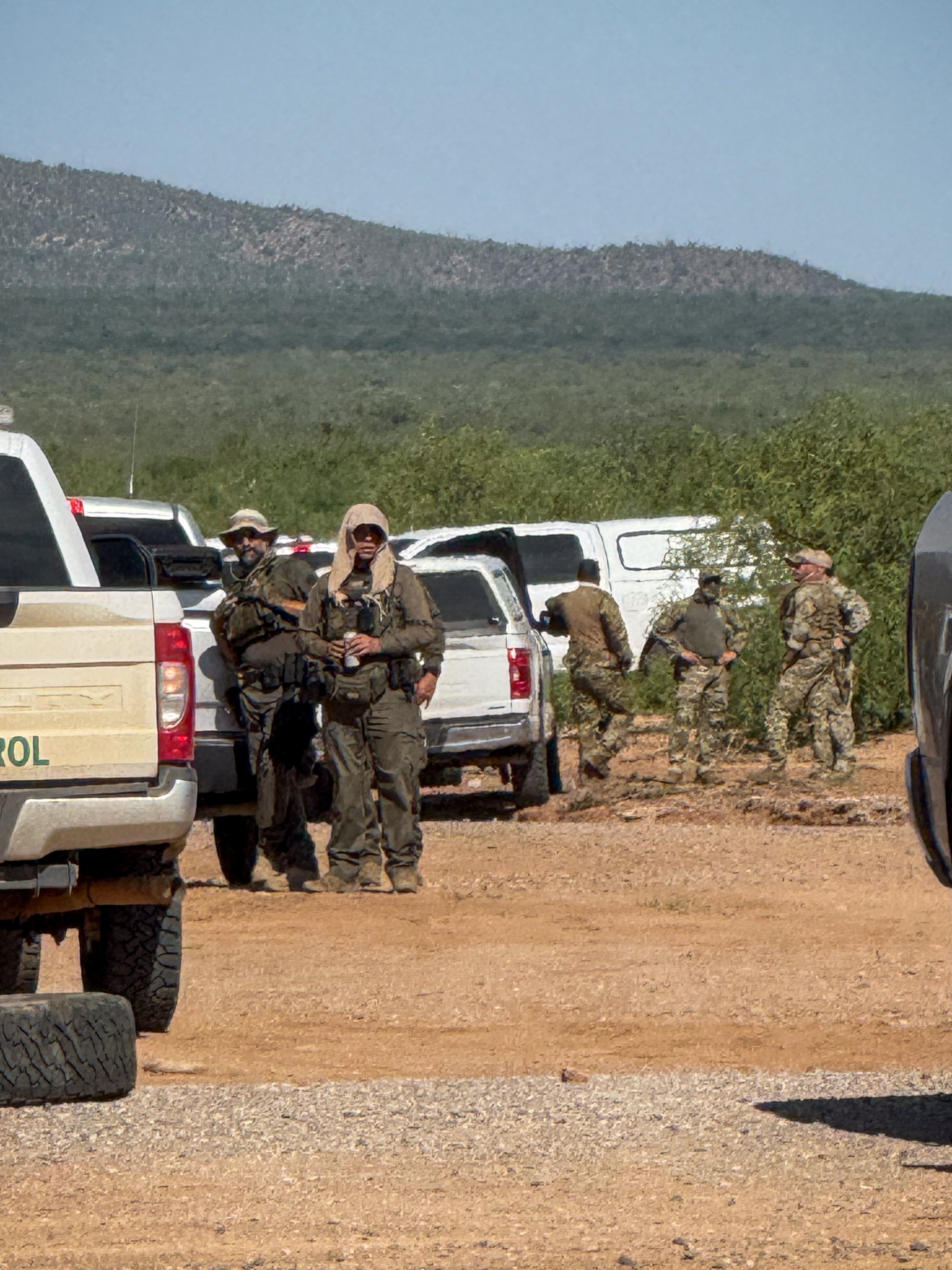 Border Patrol agents in tactical gear stand near parked white and green trucks in a desert landscape.