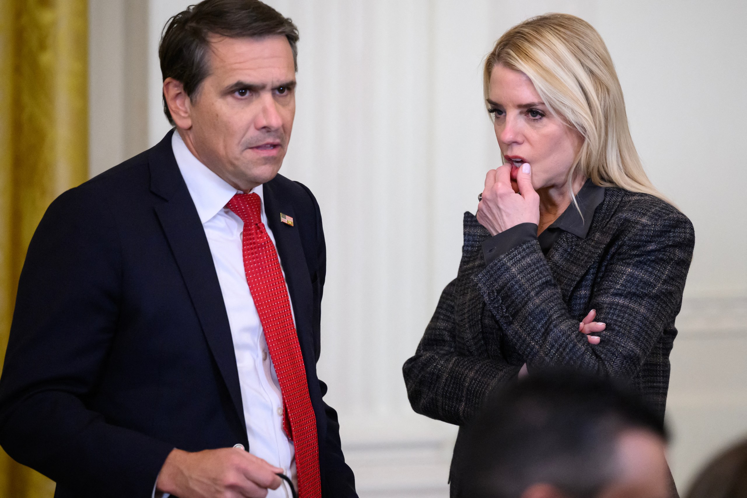 A man in a dark suit and red tie speaks with a woman in a plaid jacket who is holding her bottom lip during an event at the White House.