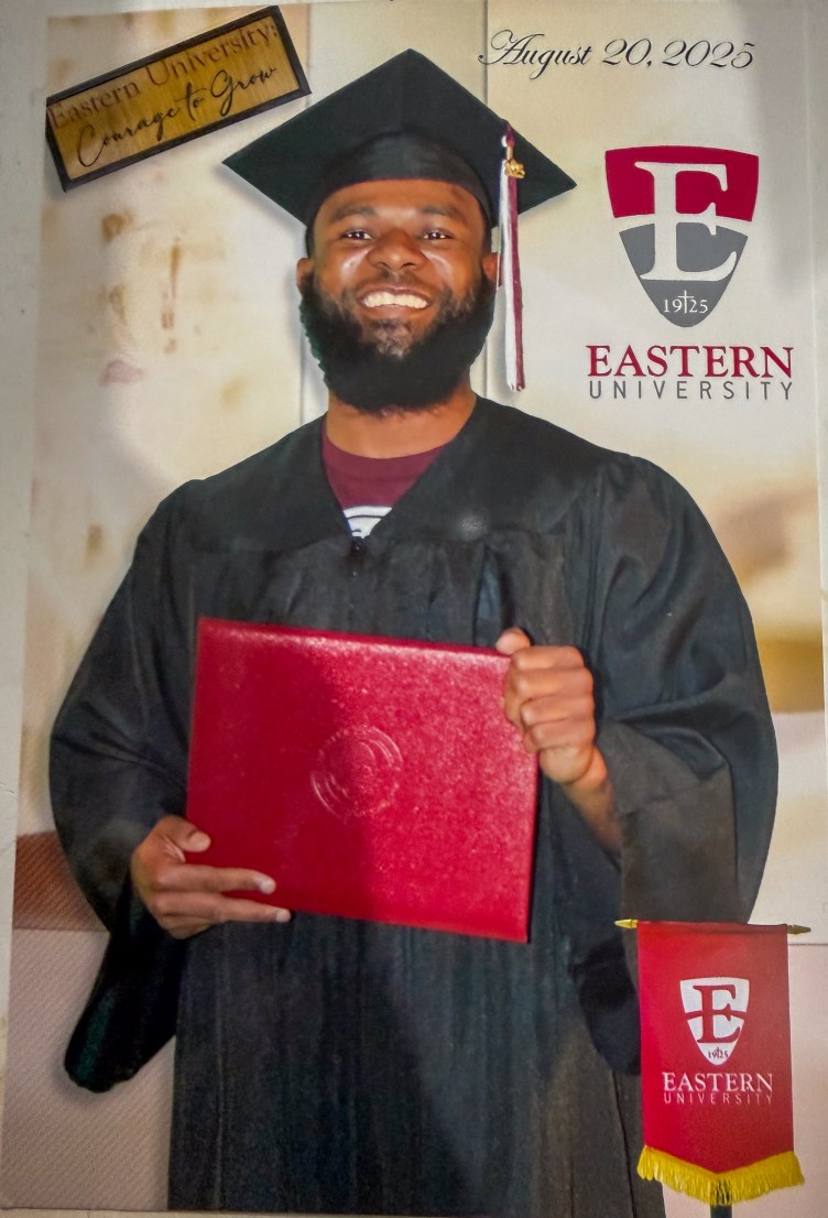 A young bearded man in a cap and gown flashes a broad smile while holding a diploma in both hands in front of his torso.