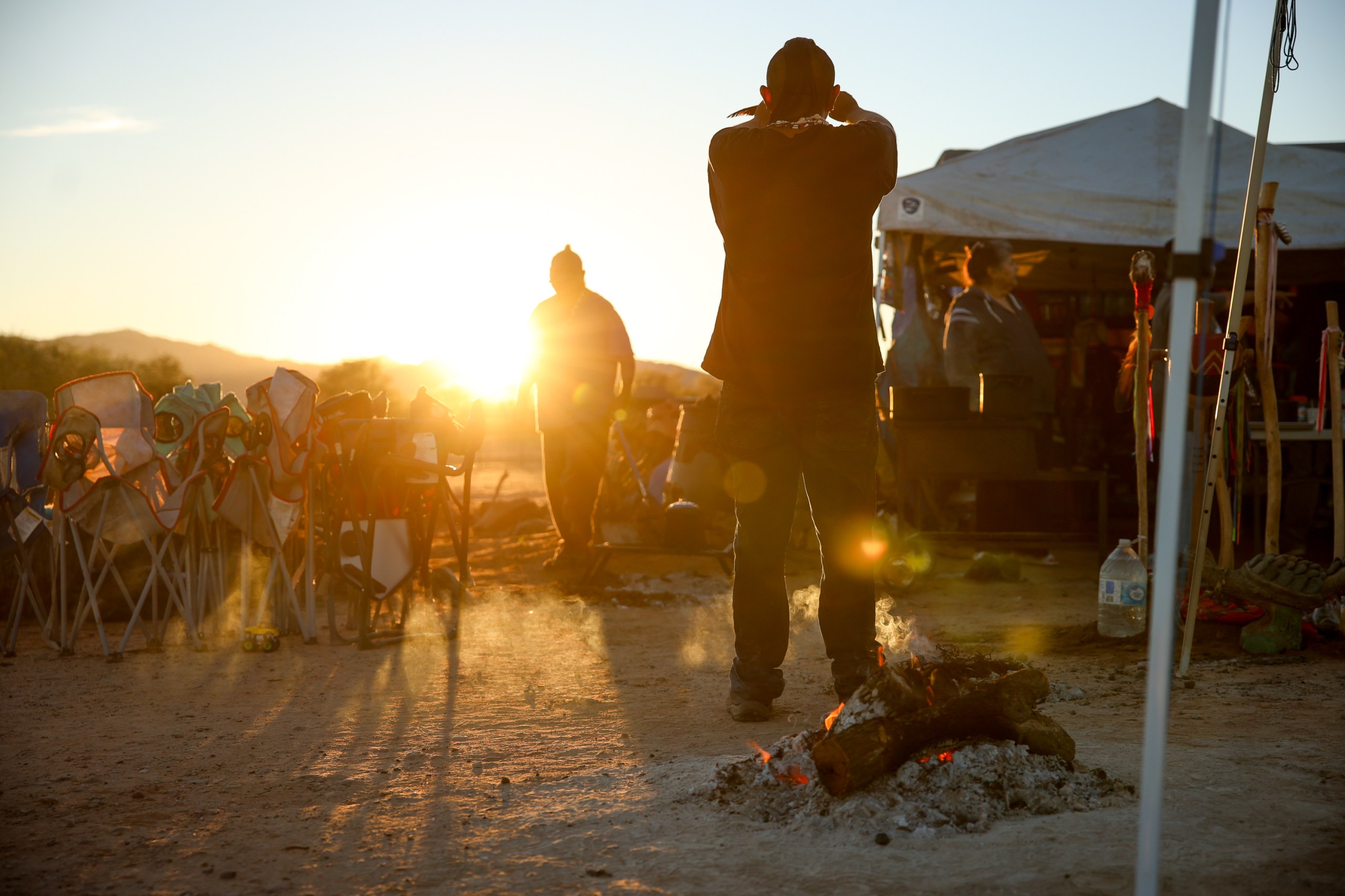 Two individuals standing in a campsite are silhouetted against a rising sun, while a ceremonial fire burns in the foreground.