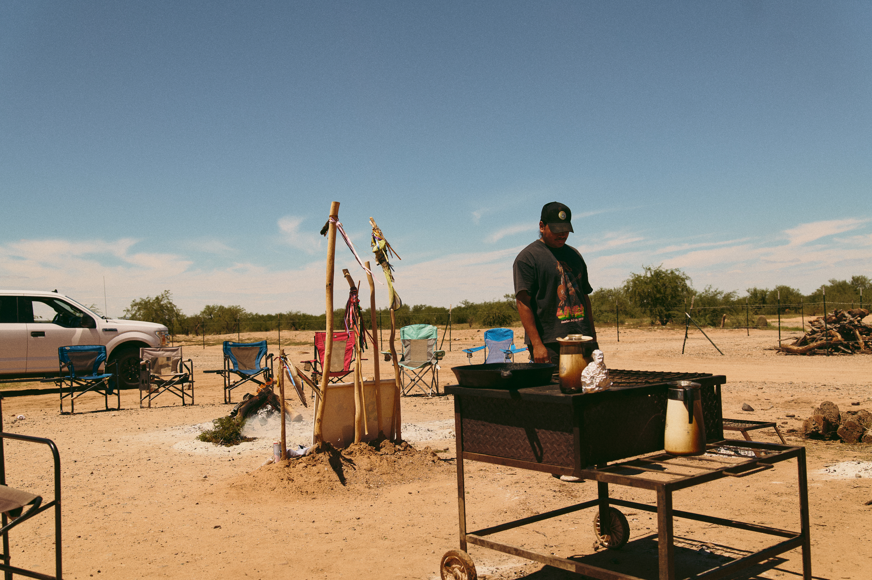 An Indigenous person stands near a burning fire surrounded by camping chairs in a desert landscape campsite.