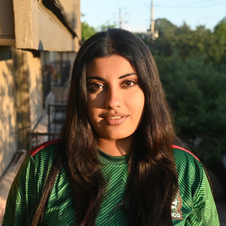 A woman with long black hair wears a green top and smiles at the camera. Her face is dramatically lit and she is in front of a building and out-of-focus trees.