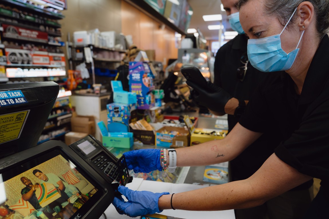 A person wearing blue gloves removes an overlay from a card reading machine at a convenience store checkout counter.