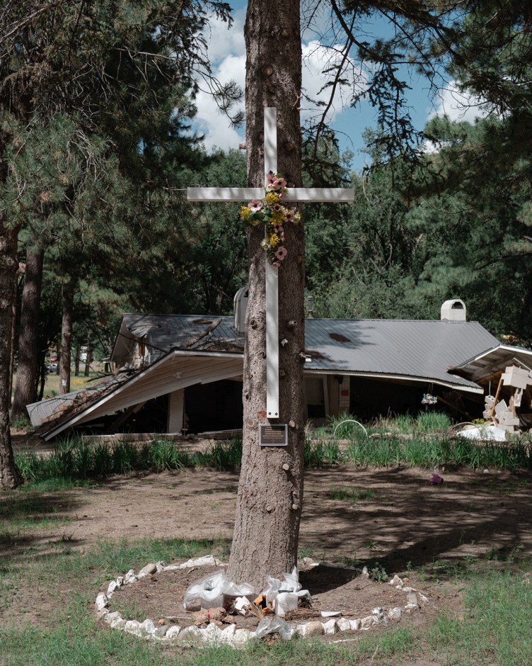 A wooden cross decorated with flowers stands attached to a tree in front of a collapsed building in a wooded area.