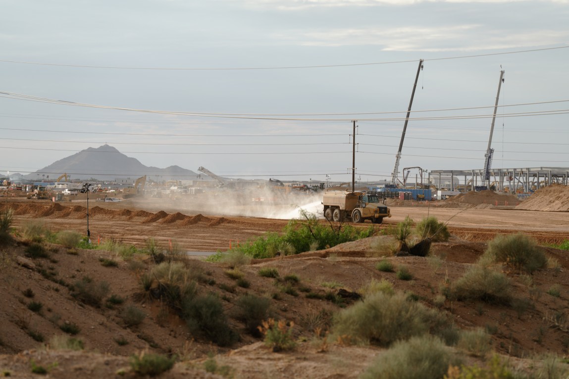 A truck sprays water across a dusty, wide-open excavation site with cranes and mountains visible in the background.