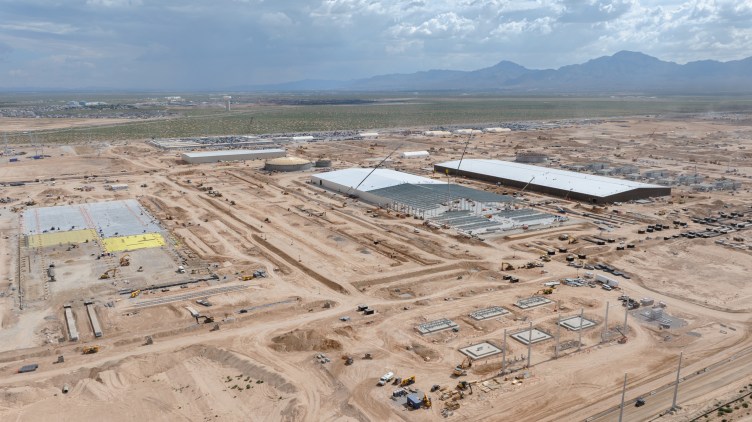 An aerial view shows a massive industrial construction site with large building foundations and steel frameworks set across a wide desert landscape.